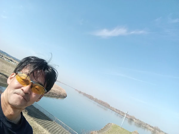 利根川を背景に、黄色のスポーツサングラスをかけた男性が自撮りしている様子。晴天の屋外でアクティビティを楽しんでいる雰囲気です。
