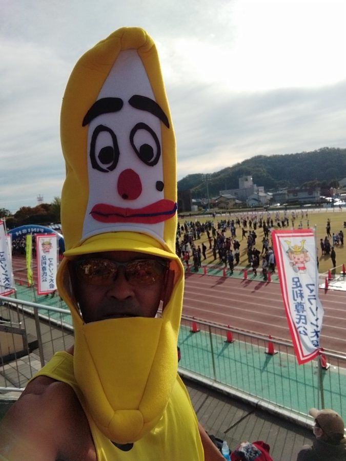 スタート前、陸上競技場のスタンドで撮ったバナナの被り物の自撮り。トラックと開会式に集まるランナーが見える