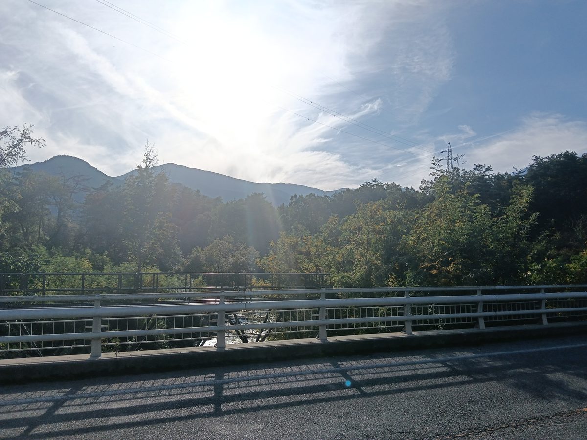 橋の上から、日差しを受けた山並みと川沿いの木々を見る