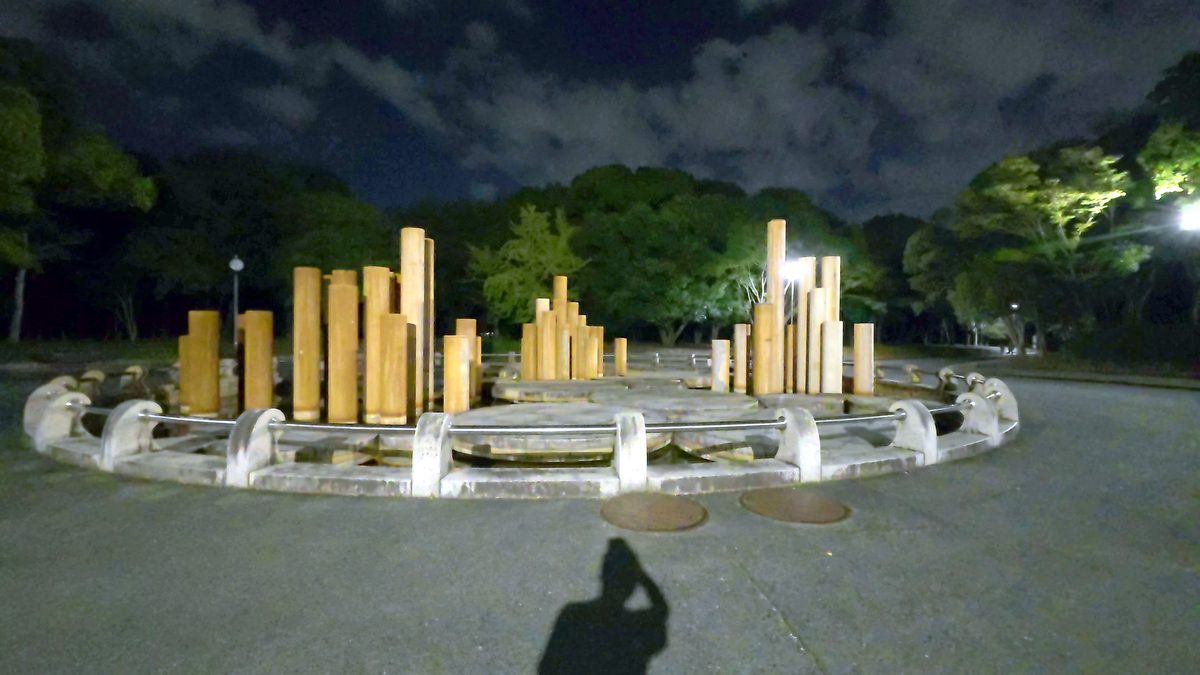 夜の公園の広場。円形の台の上に、高さの違う木の柱がたくさん立っているモニュメントと、撮影者の影