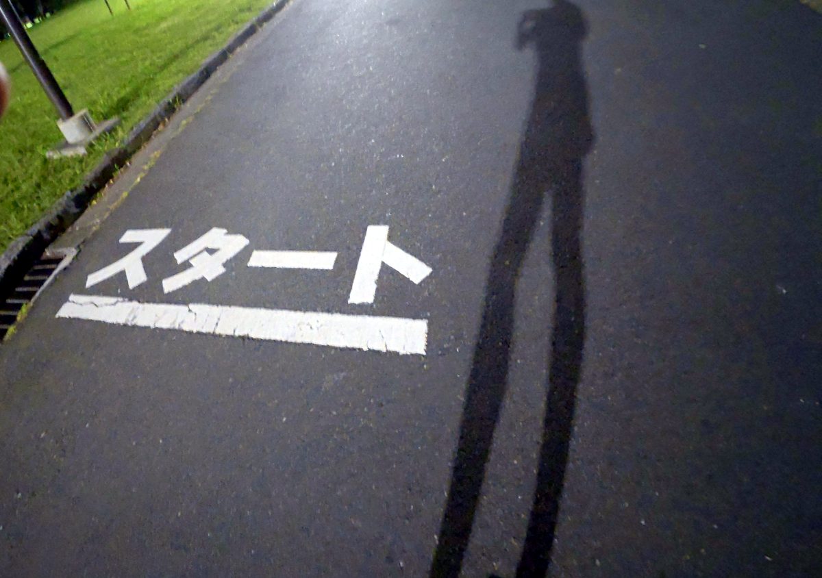 夜の公園の舗装路に白い文字で書かれた「スタート」の線と、ランナーの影